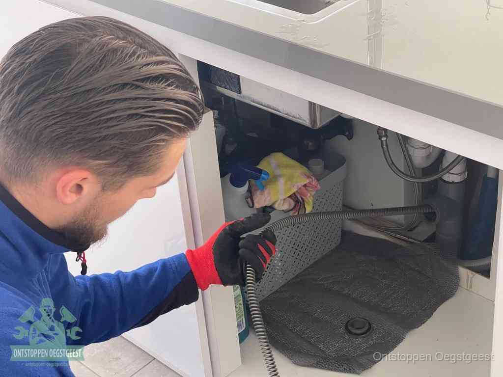 Een monteur voert een flexibele ontstoppingsveer in onder het keukenkastje, langs de sifon, om de gootsteen te ontstoppen in Oegstgeest