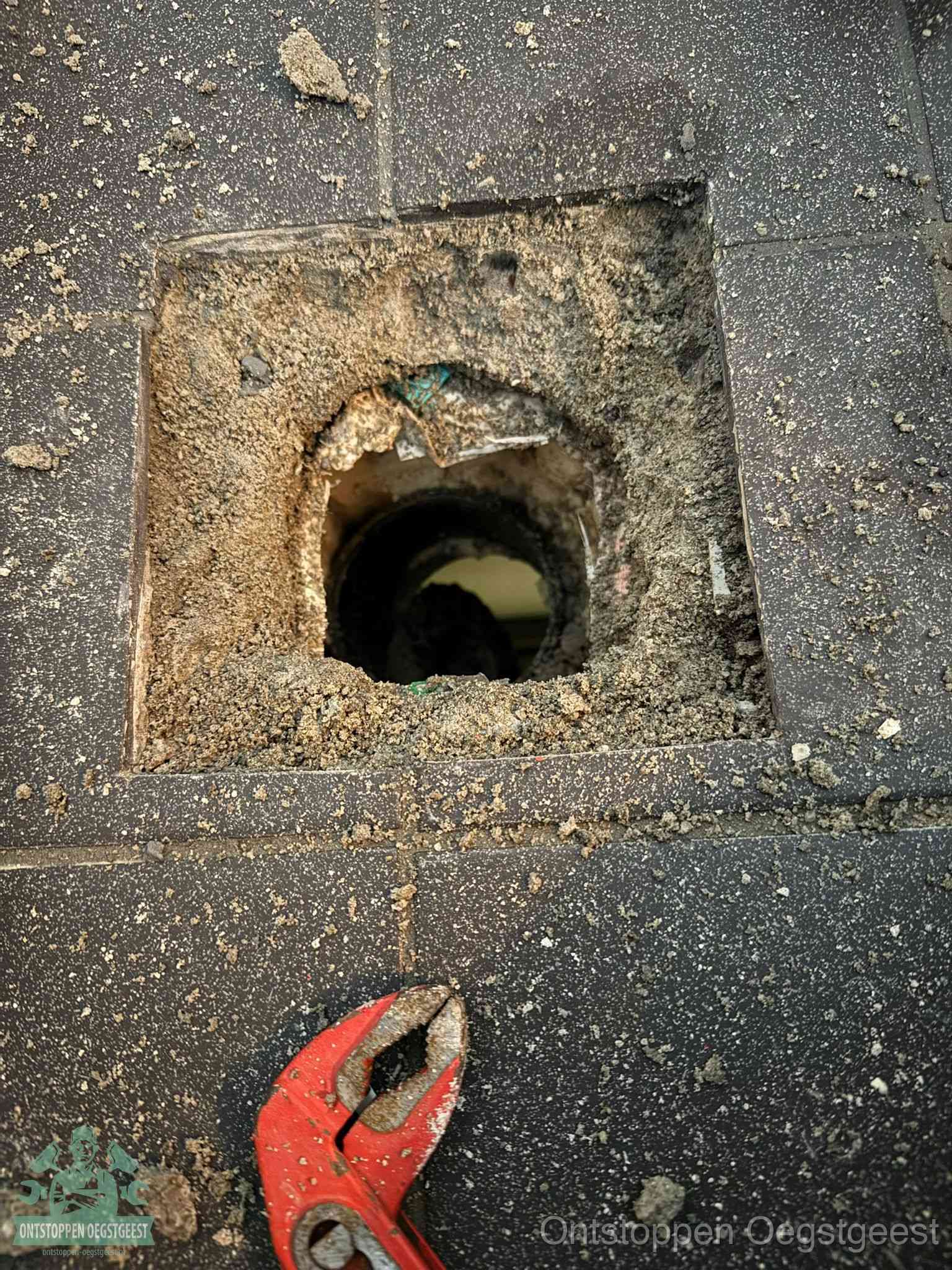 Een geopend rioolputje in een betontegelvloer met losgemaakt rooster en een zichtbare ronde afvoerbuis, het achterhalen waar de afvoerverstopping zit in Oegstgeest