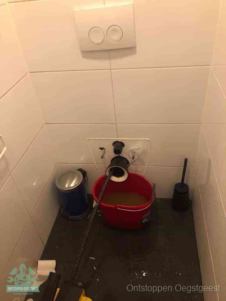 Een ontstopte wand-wc met verwijderde toiletpot waar een ontstoppingsveer in de wanduitgang verdwijnt, het achterhalen waar de verstopping zit in Oegstgeest