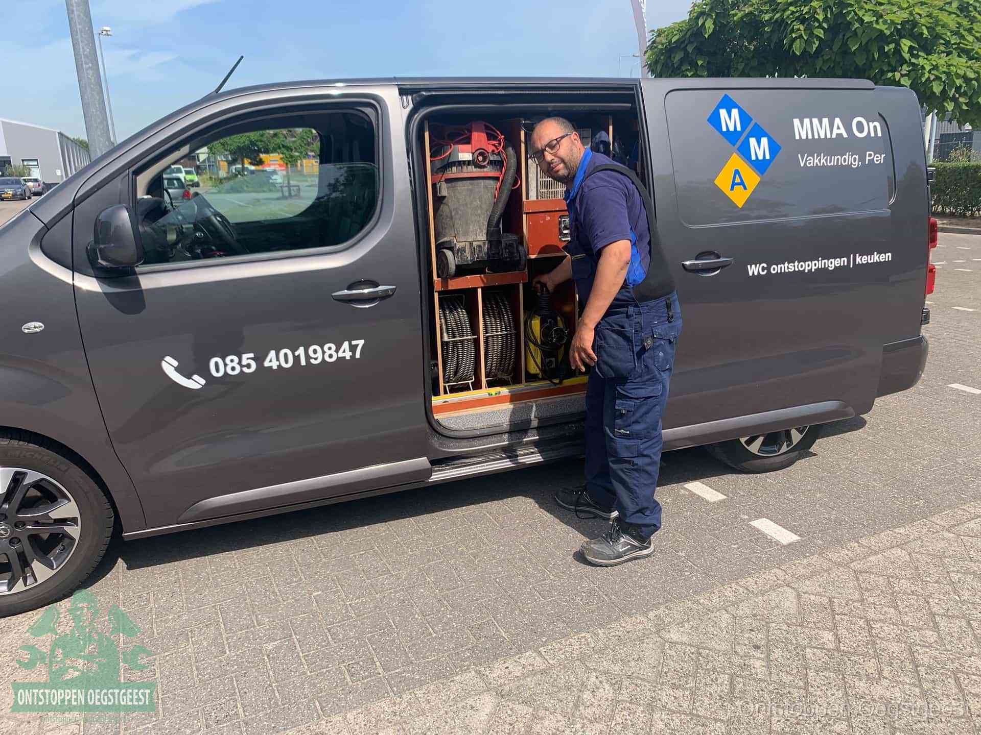 Een monteur bij de open schuifdeur van zijn bedrijfsbus op een parkeerplaats, klaar om de oorzaak van terugkerende afvoerverstoppingen aan te pakken in Oegstgeest