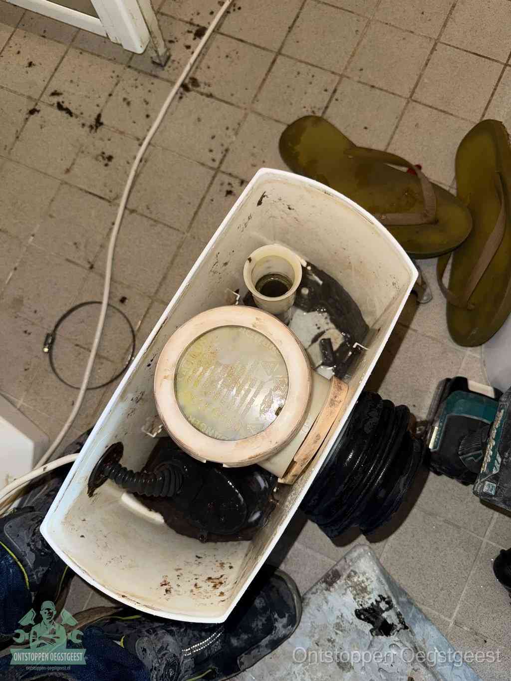 Een gedemonteerde toiletpot op de vloer met ontstoppingsveer en gereedschap ernaast, het verwijderen van een vastzittend voorwerp uit de wc in Oegstgeest
