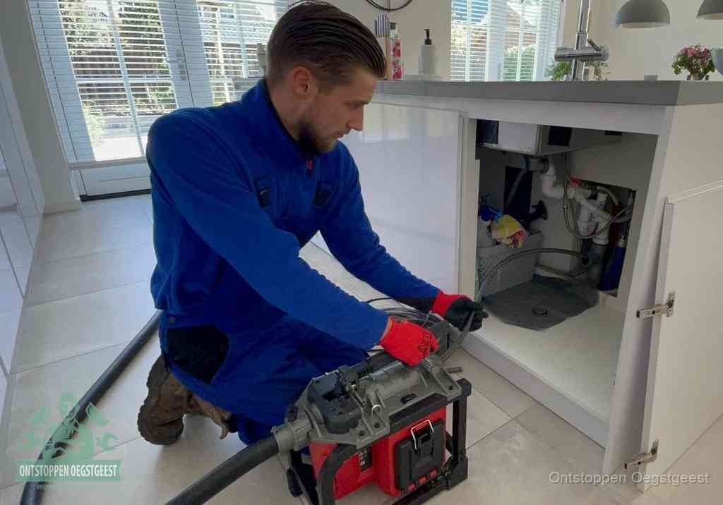 Een monteur voert een professionele elektrische ontstoppingsmachine in de afvoer onder de spoelbak tijdens het gootsteen ontstoppen in Oegstgeest